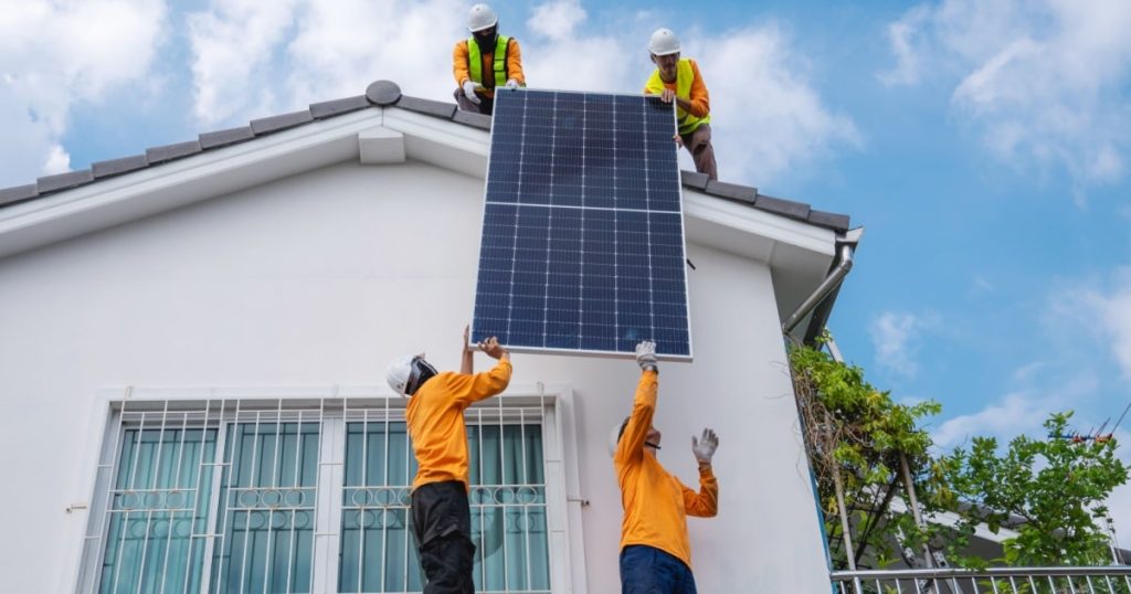 Devant une maison, deux ouvriers au sol tendent à deux ouvriers sur le toit un panneau solaire pour l'installer sur la toiture. 